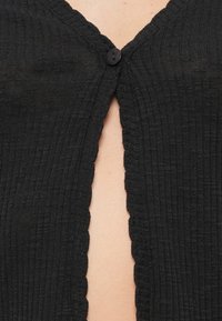 Μαύρο ριμπ ζακέτα με κούμπωμα κουμπιών στη λαιμόκοψη και κυματιστά άκρα κατά μήκος του ανοίγματος, που αναδεικνύουν υφή υφάσματος.