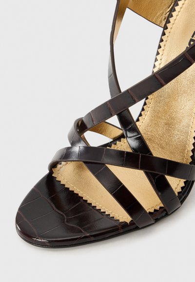 Dark brown heeled sandal with a crocodile texture. Features crisscrossed straps and a gold-toned insole with zigzag edge detailing.
