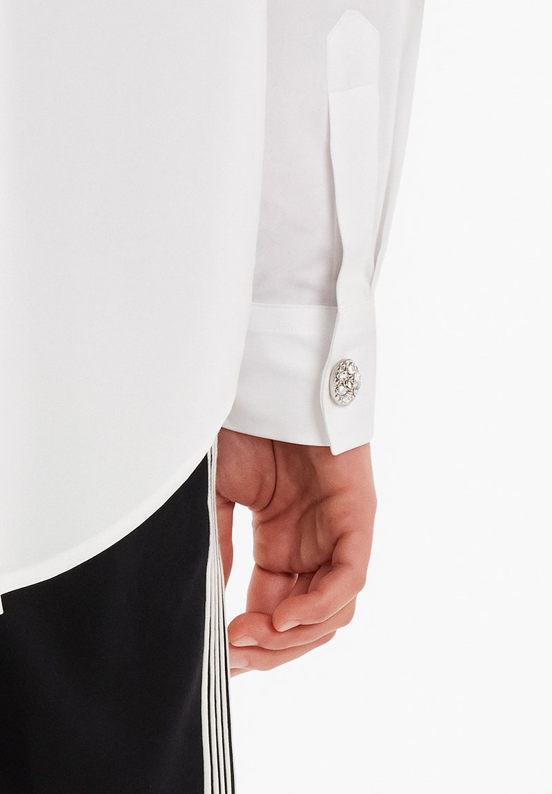 Hvid skjorte med glat tekstur, der har en buet kant og manchet med knap prydet med en dekorativ ædelstensknap. Sorte bukser med frynser.