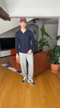 Νέος άνδρας με κόκκινο καπέλο, μπλε πουλόβερ, γκρι φόρμα και αθλητικά παπούτσια, στέκεται σε ένα σαλόνι με καφέ καναπέ και μεγάλο γλαστράκι.