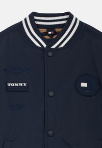 Chaqueta bomber azul marino con un cuello acanalado a rayas, logotipos bordados y cierre de botones a presión. Tejido suave con un ligero brillo.