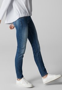 Hubené modré džíny s různými odstíny, mírně odřené na stehnech, spárované s bílými teniskami a šedým pruhovaným topem.