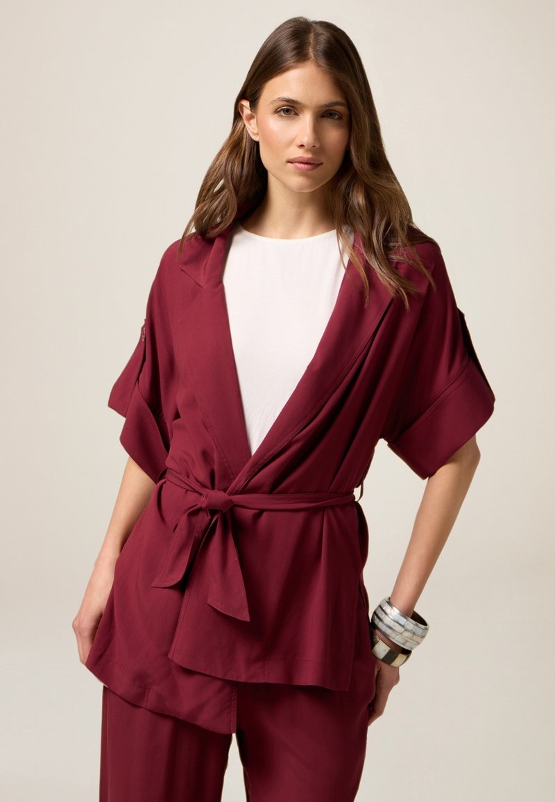 Burgundin värinen kimono-tyylinen takki, jossa on leveät hihat ja sidottu vyötärö, yhdistettynä valkoiseen paitaan ja kerroksellisiin rannekoruihin. Takissa on pehmeä kangas.
