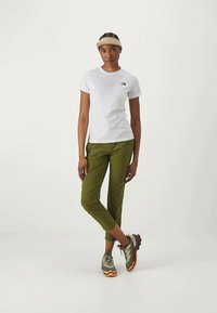 Femeie în picioare, purtând o vizieră bej, un tricou alb cu mânecă scurtă, pantaloni scurți oliv și pantofi de drumeție verzi cu tălpi portocalii.