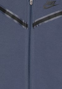 Chaqueta con cremallera de Nike en tejido azul oscuro con detalles en negro, que presenta un cuello elevado, colocación del logo y un acabado suave y texturizado.
