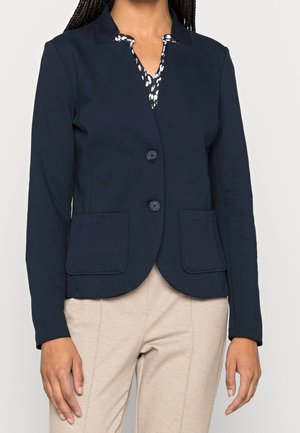 Kvinde iført en mørkeblå blazer med to knapper og påsatte lommer over en sort-hvid mønstret bluse, kombineret med beige bukser.