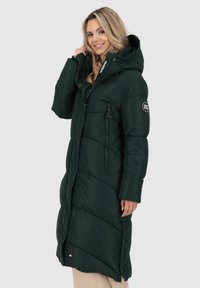 Giacca puffer verde con cappuccio, caratterizzata da una texture trapuntata, tasche laterali e una zip frontale. Indossata da una modella con capelli lunghi.