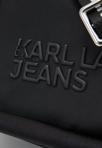 Fekete táska texturált külsővel, dombornyomott "KARL LA JEANS" logóval és ezüst színű kiegészítőkkel, minimalista dizájnnal.