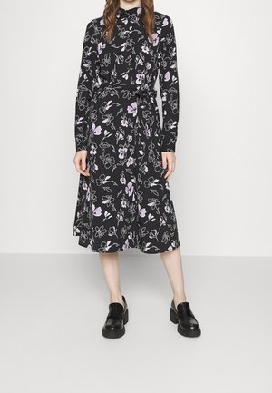 Frau trägt ein schwarzes knielanges Kleid mit lila und weißen Blumenmustern und langen Ärmeln, kombiniert mit schwarzen klobigen Loafers.