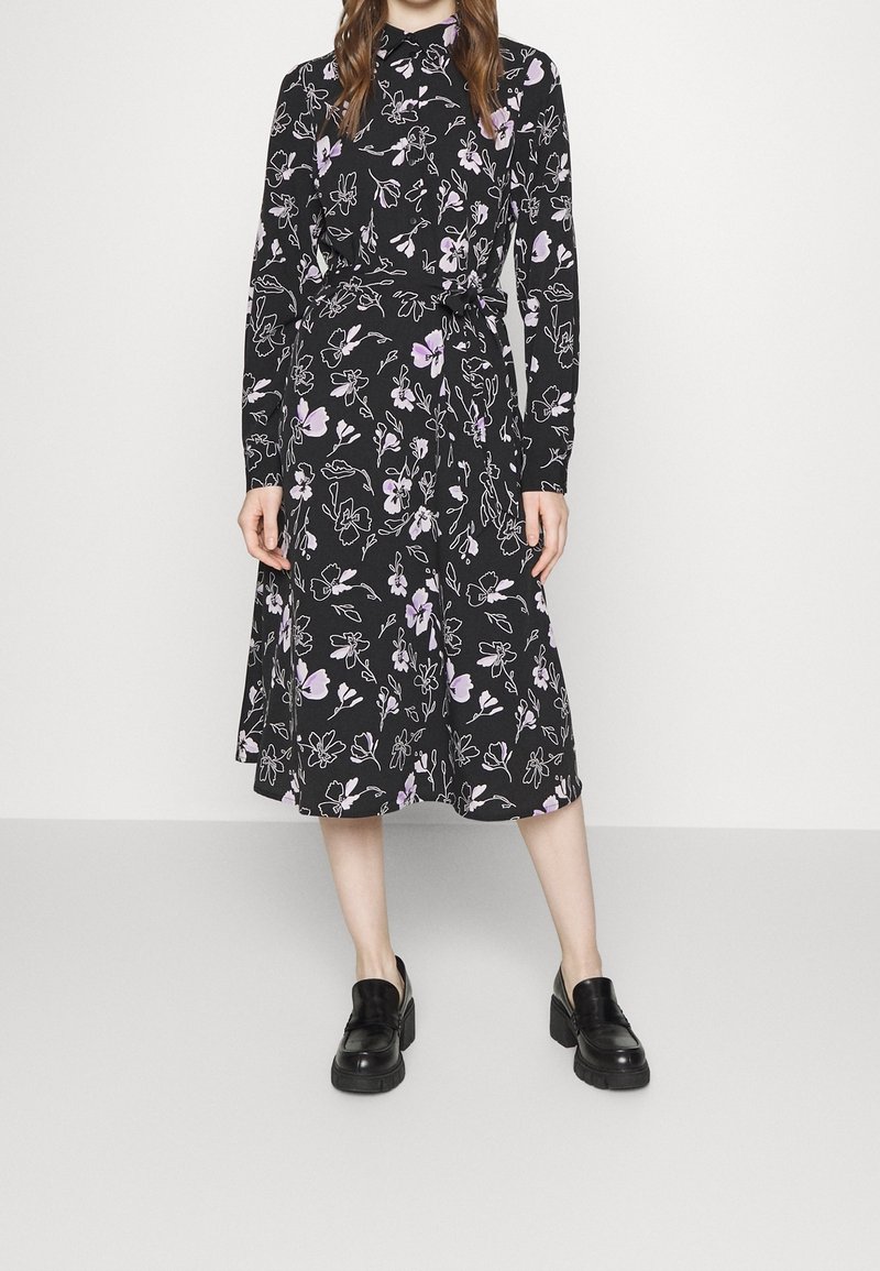 Femme portant une robe noire longueur genou avec des motifs floraux violets et blancs et des manches longues, associée à des mocassins noirs épais.