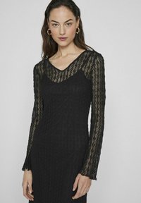 Melnās mežģīņu kleitas ar garām, nedaudz izplestām piedurknēm un V-veida kakla izgriezumu. Audumam ir teksturēts raksts, kas uzlabo tā vizuālo dziļumu.