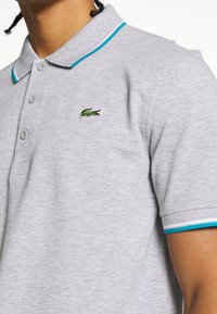 Grå polo skjorta med en strukturerad yta. Har turkosa accenter på kragen och ärmarna, samt en liten grön krokodil-logga på bröstet.