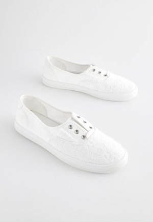 Bijele tenisice od čipke s teksturiranim cvjetnim uzorkom, rastezljivim bočnim stranama i srebrnim rupama za pertle. Gumeni potplati pružaju prianjanje.