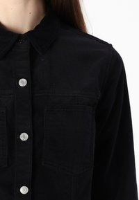 Black denim shirt with a button-down collar, silver button closures, and two chest pockets. Soft texture and a relaxed fit.