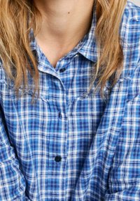 Blue plaid button-up shirt with a collar. Made of fabric with a textured appearance. Features dark buttons and contrasting white lines.