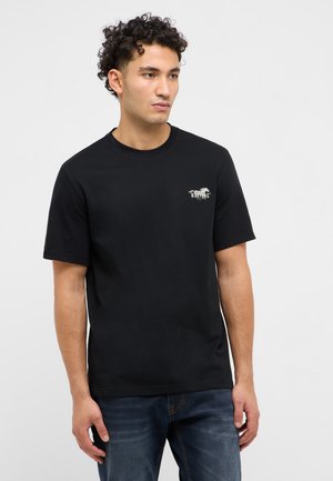 Młody mężczyzna o kręconych włosach, ubrany w czarny t-shirt Mustang i niebieskie jeansy, stojący na tle jasnego, jednolitego tła.