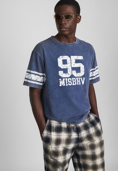 Blue cotton T-shirt with distressed neckline, featuring white graphic "95 MISBHV" and textured stripes on sleeves. Paired with plaid pants.