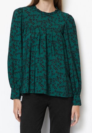 Kobieta ubrana w bluzkę w kolorze zielono-niebieskim z czarnym kwiatowym wzorem na długim rękawie i czarne spodnie, stojąca na białym tle.