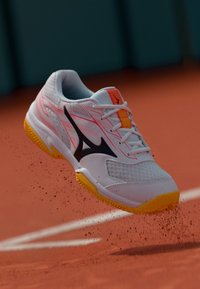 Białe buty sportowe z różowymi akcentami, czarnym logo i żółtą podeszwą. Posiadają siateczkową cholewkę i charakterystyczny design, zawieszone nad powierzchnią boiska.