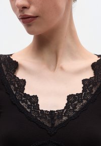 Μαύρο τοπ με βαθύ V-λαιμό με φλοράλ δαντέλα. Η δαντέλα έχει περίτεχνα σχέδια και συμπληρώνει το λείο, εφαρμοστό ύφασμα.