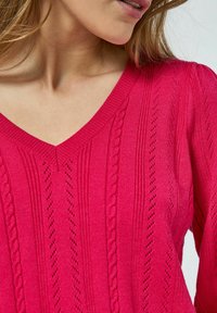 Jasnoróżowy sweter z dzianiny, z dekoltem w serek, prążkowaną teksturą oraz szczegółowymi wzorami z motywem warkoczy i dziurek.