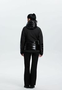 Sort isoleret jakke med en blank hætte og taljepanel, der har en mat underkrop og lange sorte bukser. Minimalistiske designelementer.