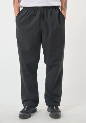 Mann trägt lockere schwarze Hose mit elastischem Bund und schwarze Sneakers, steht mit den Händen in den Taschen.