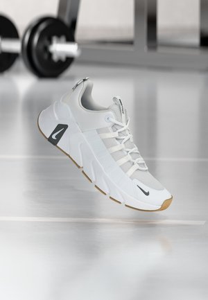 Witte en grijze sportschoen met gumzool en zwart logo, zwevend boven een reflecterende vloer nabij fitnessapparatuur.