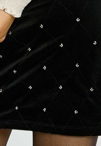 Falda de terciopelo negro con un patrón acolchado en forma de diamante, acentuada con pequeñas decoraciones plateadas distribuidas de manera uniforme por toda la tela.