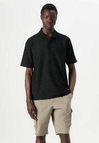 Svart kortermet polo skjorte med krage-design, laget av mykt stoff. Brukt med beige shorts som har sidelommer.