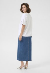 T-shirt blanc avec des détails cloutés sur les manches, associé à une longue jupe en denim bleu et des sandales à talons blancs, vu de dos.