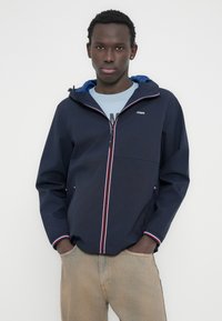 Ναυτικό μπουφάν με κουκούλα, με αντιθετικές λεπτομέρειες φερμουάρ σε κόκκινο και άσπρο χρώμα· ελαφρύ ύφασμα· διαθέτει πλαϊνές τσέπες και κουκούλα με μπλε φόδρα.