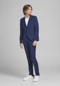 Costume ajusté bleu marine avec un revers cranté, deux boutons et des poches avant, associé à une chemise blanche à col et des baskets blanches.