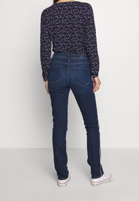 Tmavě modré džíny z denimu s rovnými nohavicemi, s kapsami na zadní straně a bočními pruhy v červené a bílé barvě. Viditelná je také vzorovaná košile s dlouhými rukávy.