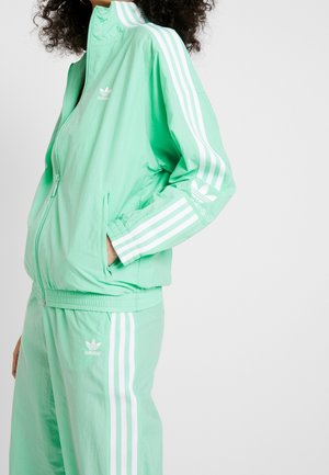 Femme portant un survêtement Adidas vert menthe avec des détails à trois bandes blanches et des logos sur la manche de la veste, la poitrine et le pantalon.