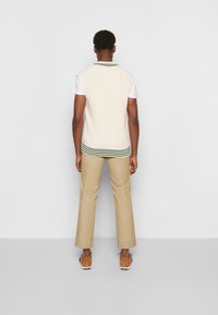 Gestrickte cremefarbene Pulloverweste mit grün gestreiftem Saum über einem weißen T-Shirt, kombiniert mit beigen Hosen und braunen Schuhen, Rückansicht.