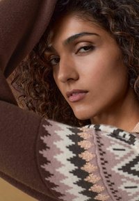 Pull en maille avec des motifs géométriques dans des nuances de marron, rose et blanc cassé. Présente un large poignet et un tissu texturé le long de la manche.