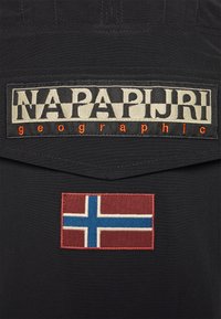 Tessuto nero con un'importante etichetta "NAPAPIJRI geographic" crema e arancione e una bandiera norvegese ricamata in rosso, bianco e blu.