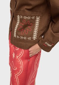 Hnedá bunda s bielymi a červenými vyšívanými vzormi na vrecku a manžete, nosená cez červenú vzorovanú sukňu, ruka vrecku.