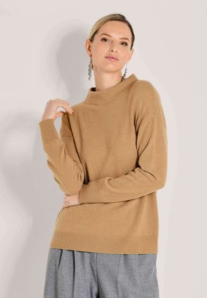 Vrouw met blond haar draagt een beige trui en grijze broek, poseert met een gebogen arm dicht bij haar gezicht tegen een lichte achtergrond.