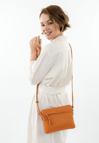 Borsa a tracolla in pelle arancione con superficie testurizzata, dotata di tasca frontale con zip e tracolla regolabile. Dimensioni compatte, per un facile trasporto.