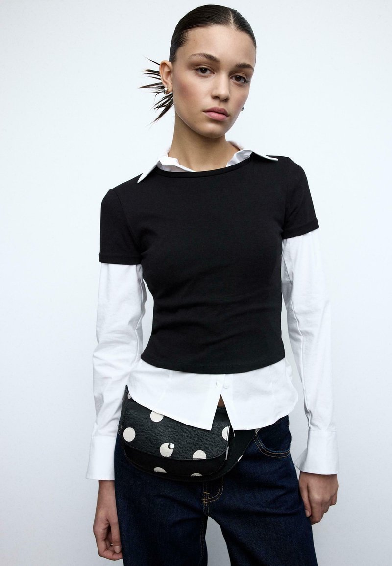 Haut noir ajusté superposé à une chemise blanche à boutons, accompagné d'un jean bleu et d'un sac bandoulière à pois. Design lisse et décontracté.