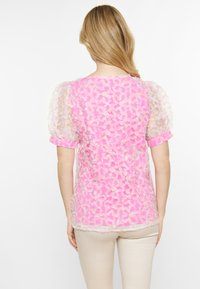 Lyserøde, gennemsigtige bluse med struktureret blomstermønster, puffede kortærmer og rund udskæring. Lyse detaljer på ærmerne fremhæver designet.