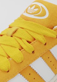 Yellow suede sneaker featuring a textured finish, white leather stripes, and matching yellow laces. The tongue displays a white logo accent.