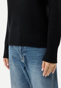 Svart stickad tröja med ribbade muddar och fåll, kombinerad med ljusblå denimjeans. Fokus på textur och passform för en avslappnad look.