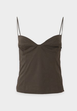 Une femme porte un caraco olive, un pantalon noir à jambes larges et des sandales sur un fond uni et clair.