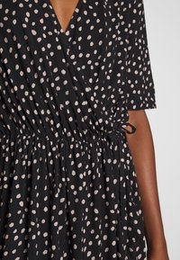 Robe noire enroulée avec des pois beige clair, manches courtes et taille froncée avec un détail à nouer. Tissu doux et léger.