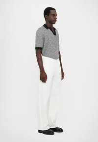 Polo din tricot gri cu guler și mansete negre, asortat cu pantaloni largi albi. Modelul poartă bocanci negri masivi. Fundal simplu.