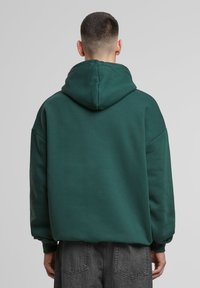Felpa verde con cappuccio, dal taglio morbido e rilassato, con spalle scese e polsini a costine. Il tessuto sembra morbido e liscio, con cuciture minime.