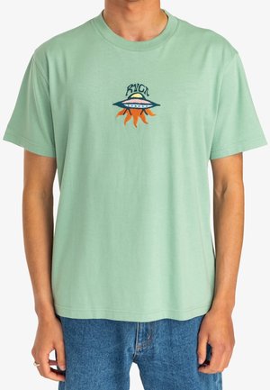 Homme portant un t-shirt vert clair avec un petit motif brodé d'un OVNI et des rayons de soleil sur la poitrine, associé à un jean bleu.
